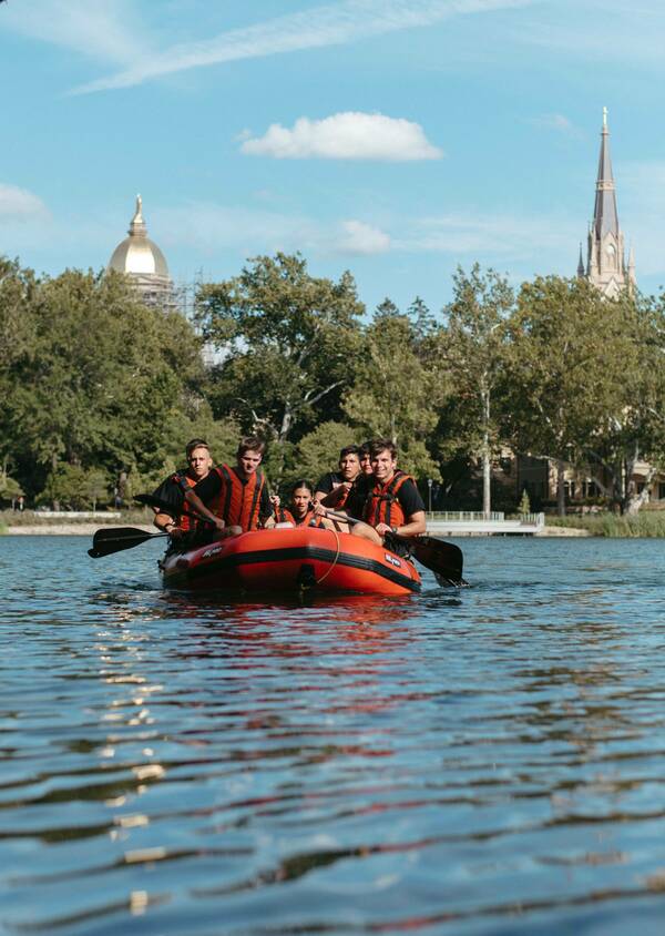 Five Army ROTC Cadets in orange row boat on Notre Dame campus lake on a sunny day.