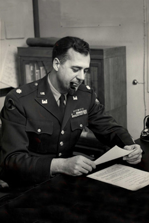 Black and white photo of LTC G. M. Cookson smoking a pipe while looking at papers at a desk with his office in background