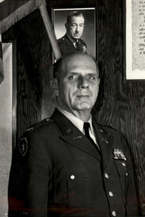 Headshot of COL E. W. Grenelle in black and white with a photo on the wall in the background