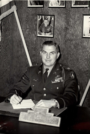 Black and white photo of COL C. J. Letzelter signing a paper at a desk with a photo on the wall in the background
