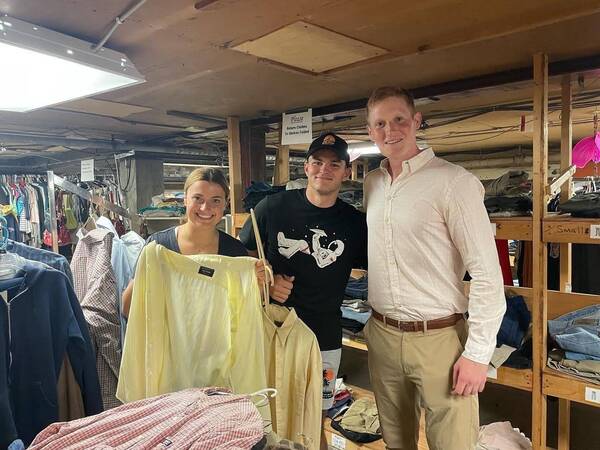 Two men and one woman pose while holding up shirts on hangars in front of racks of clothes