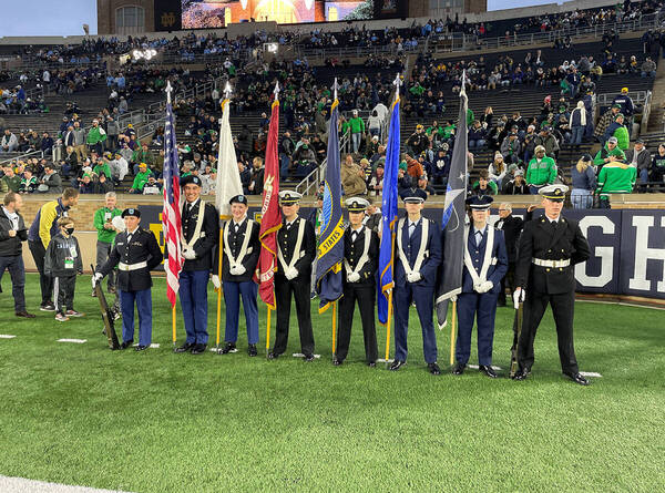 Colorgaurd composed of 8 people stand in a line on the Notre Dame football field holding various flags 