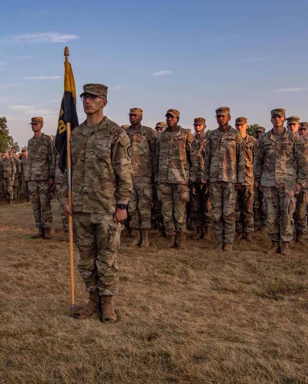 Male cadet stands stoically holding our flag in front of several other cadets who stand in attention