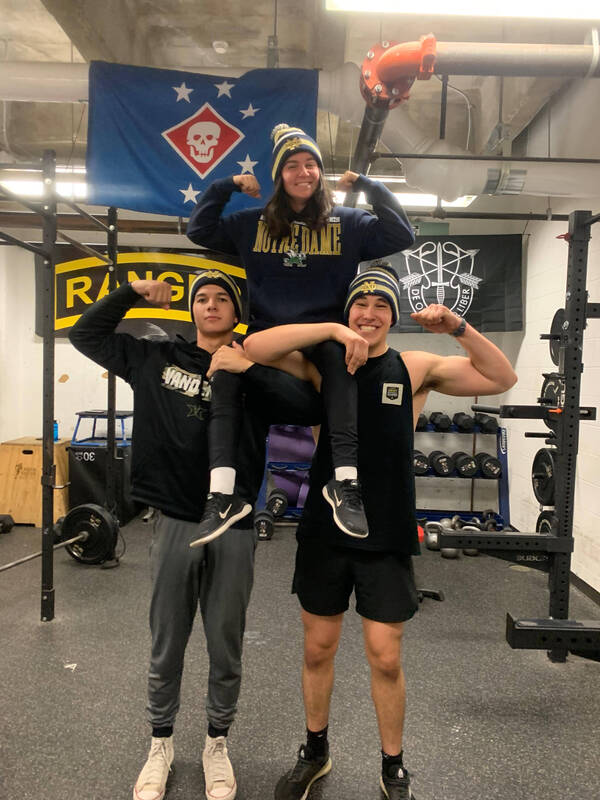 Two male cadets lift female cadet on their shoulders, while they flex in the workout gym